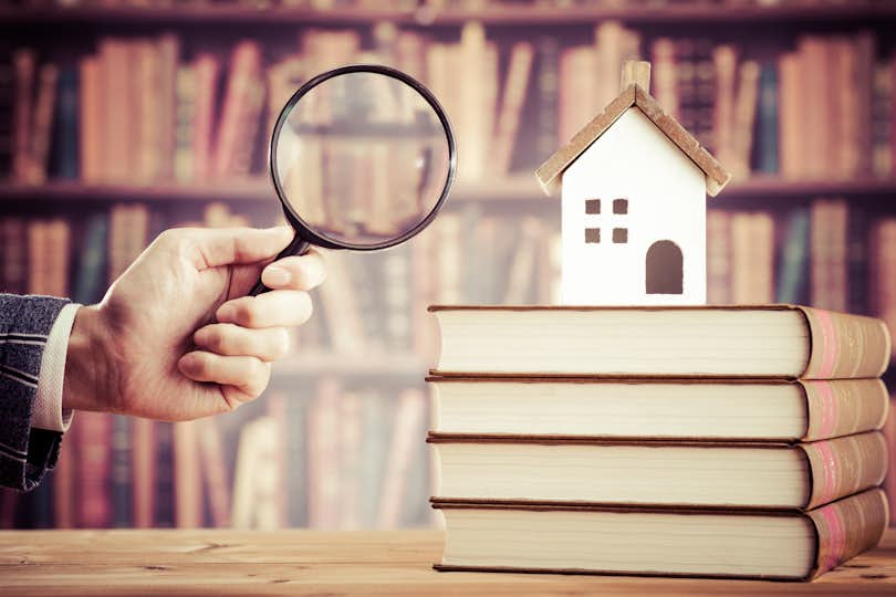 small model home on a stack of books with a hand holding a magnifying glass in the foreground