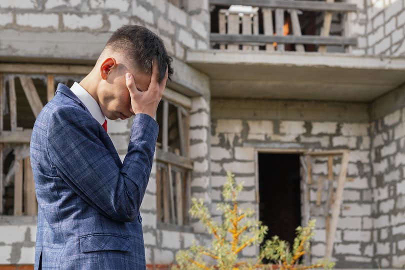 a man in a suit stands in front of an empty house in disrepair, holding his head in his hands and looking worried
