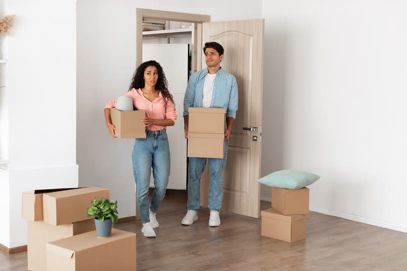 Unhappy confused millennial couple homeowners holding cardboard boxes walking in new house.