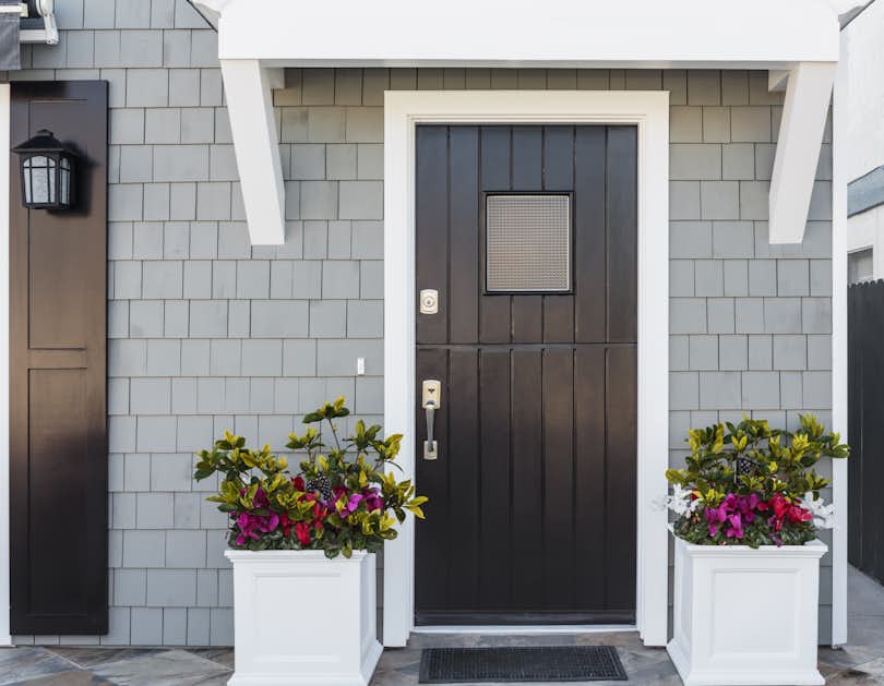 Horizontal image of black front door made of vertical wood boards, with a window, framed by two planters, gray shingles, and a door mat.